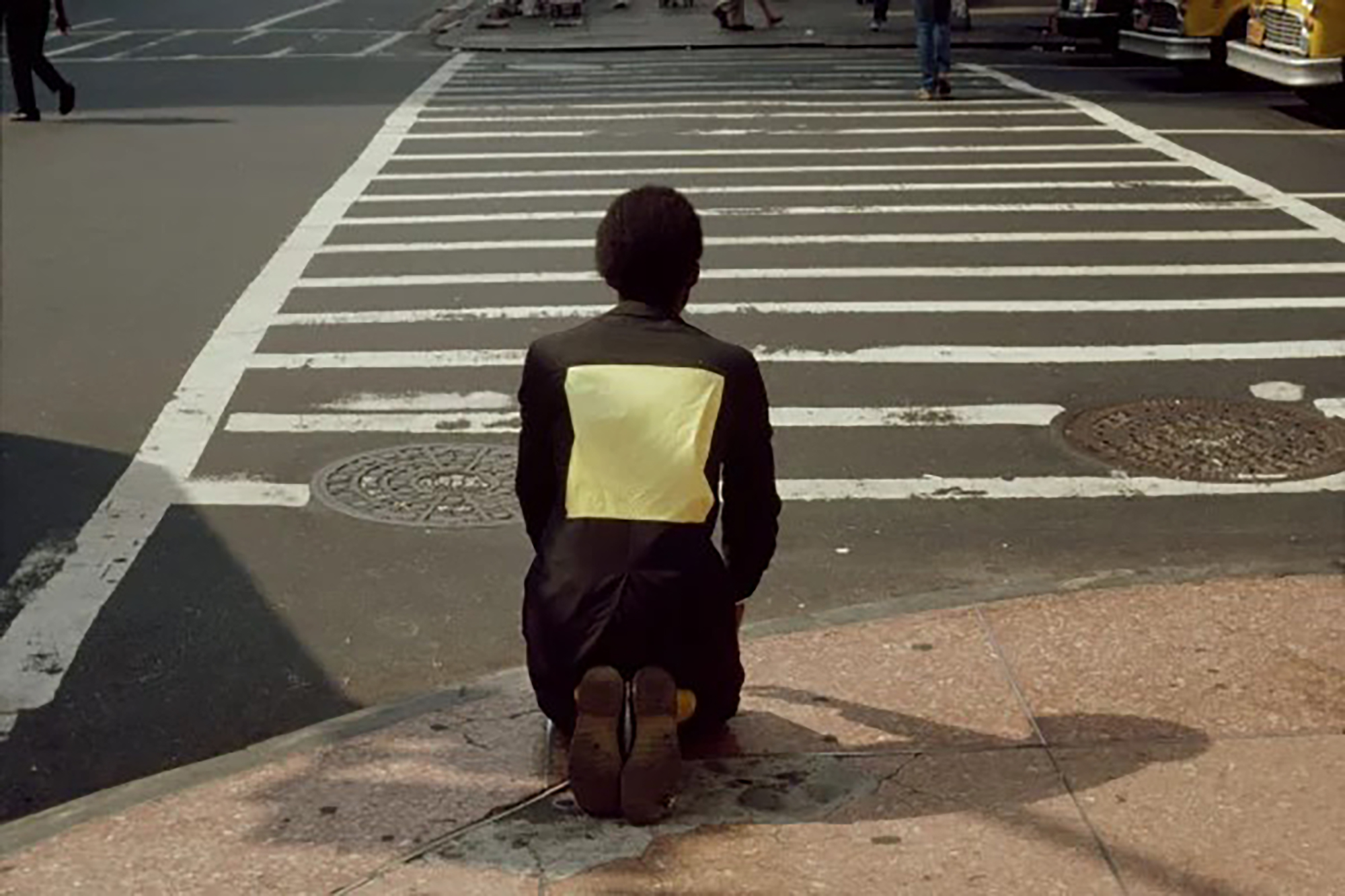 A man facing away from the camera and kneeling on a sidewalk at the entrance to a crosswalk.